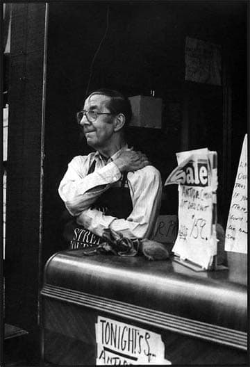 A shopkeeper in shirtsleeves and an apron leaning on a counter in his doorway beside handwritten sale signs