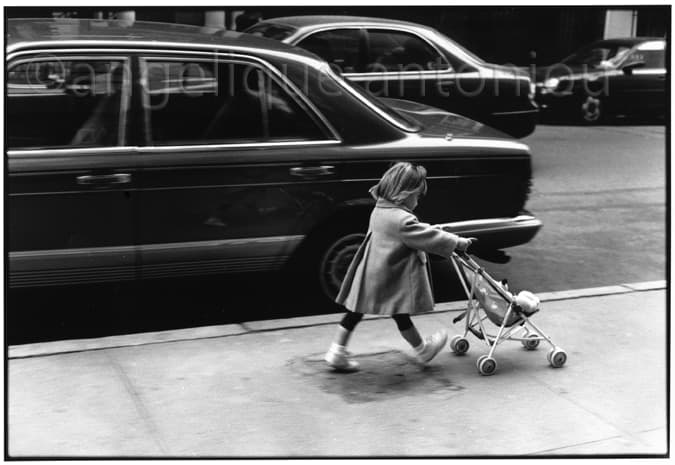 A small girl in a coat pushing a toy stroller along a city pavement past parked cars